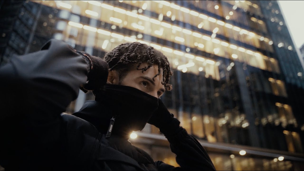 A low-angle shot of the artist looking down into the lens, gloved fists raised, framed against a glass tower rising to a vanishing point